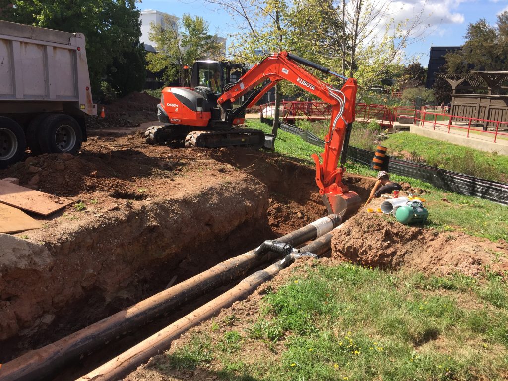 Photo of construction. A digger and a worker adjust pipes in the ground.