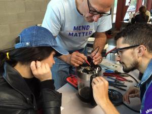A repair café technician solders the insides of an item. Two students watch his progress.